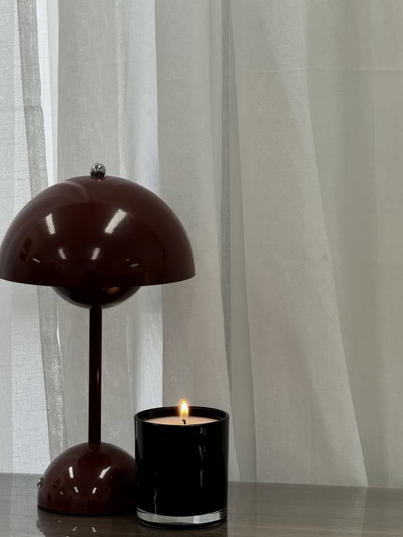An oxblood lamp and a lit candle against sheer curtains.